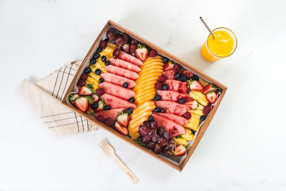 Vibrant fresh fruit platter with sliced watermelon, cantaloupe, pineapple, grapes, strawberries, blueberries, and a glass of orange juice.