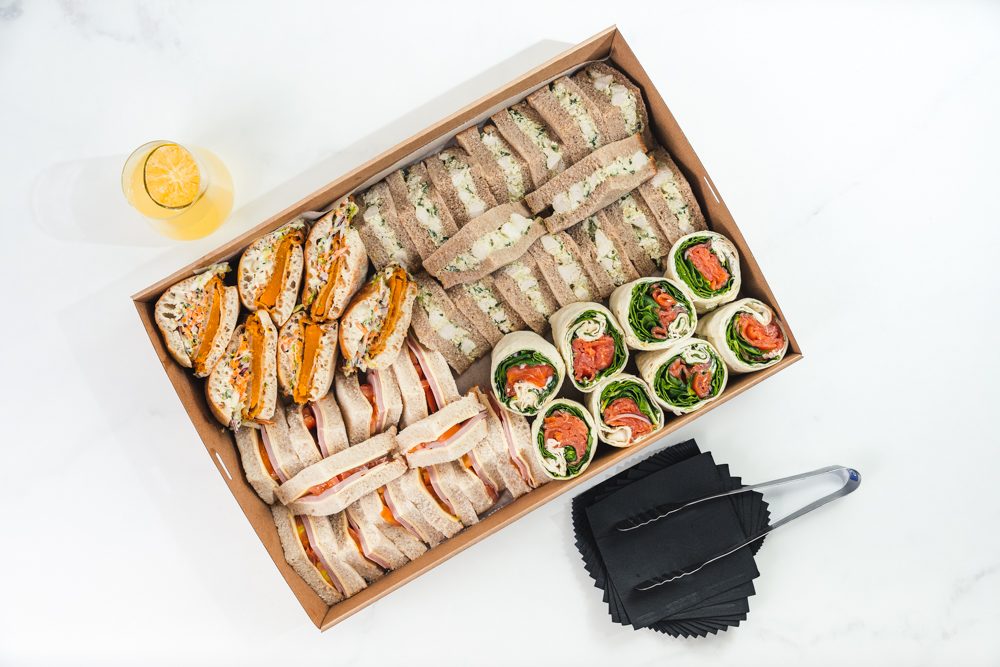 Assorted gourmet sandwich platter with wraps, mini rolls, and finger sandwiches arranged in a catering box on a white background.
